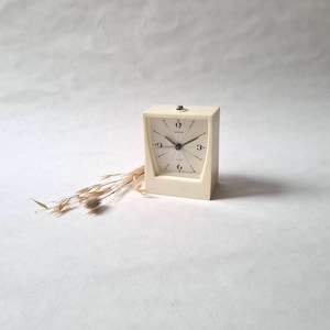 Può includere: Un orologio da sveglia vintage bianco con quadrante nero e lancette argentate. L'orologio è del marchio "Dugena" e ha una forma quadrata.