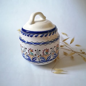 Peut inclure: Un pot en céramique blanche avec un couvercle bleu et blanc et un motif floral coloré. Le pot a une poignée et est décoré de peinture bleue, orange, jaune et verte.
