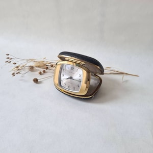 Op de afbeelding: Een gouden wekker met een witte wijzerplaat en zwarte wijzers. De klok zit in een zwarte leren koffer en staat op een wit oppervlak.