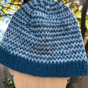 May include: A knitted beanie hat in shades of teal, blue, and white. The hat features a textured pattern and a solid teal brim. The hat is designed to fit snugly and keep the wearer warm.