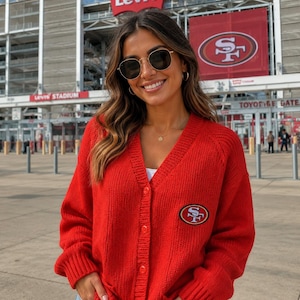 Könnte beinhalten: Eine leuchtend rote, langärmelige Strickjacke mit Knopfleiste und einem San Francisco 49ers-Logoaufnäher auf der linken Brust. Das Kleidungsstück verfügt über einen V-Ausschnitt sowie gerippte Bündchen an den Ärmeln und am Saum.