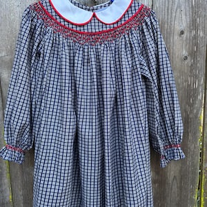 May include: A navy and white checked dress with long sleeves. It has a white Peter Pan collar edged with red, and red smocking across the chest. The cuffs are also trimmed with red. The dress is displayed on a blue hanger.