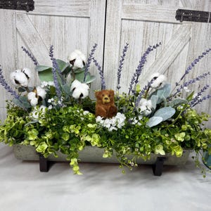 May include: A decorative floral arrangement in a rectangular container. It features cotton bolls, lavender sprigs, and greenery, with a small teddy bear figurine in the center. Two blue mason jars flank the arrangement.