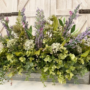 May include: A rectangular metal planter filled with a variety of faux greenery and purple lavender. The arrangement includes white flowers and various green leaves. A pink mason jar is visible in the background.
