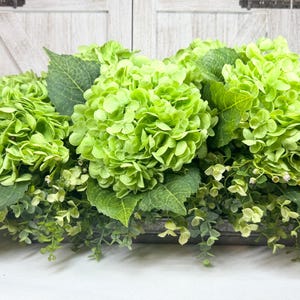 May include: A rectangular floral arrangement featuring vibrant green hydrangeas and lush greenery. The arrangement is set in a rustic, weathered metal container, creating a fresh and natural aesthetic. Perfect for home decor or event styling.