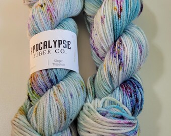 May include: Two skeins of hand-dyed yarn featuring a variegated color palette of light blue, white, lavender, and speckles of pink and gold. A white paper label wraps around the left skein with the text Apocalypse Fiber Co. Slinger, Wisconsin.
