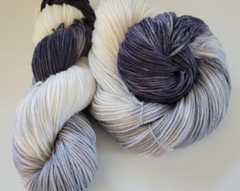 May include: Two skeins of yarn in a gradient of colors. The yarn transitions from a light gray to a darker gray, with sections of white and a deep charcoal color. The yarn is tightly wound and appears soft, suitable for knitting or crochet projects.