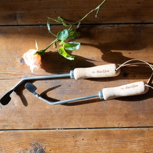 Può includere: Due attrezzi da giardinaggio con manici in legno e lame in metallo, con la scritta "The Buckhoe". Una rosa arancione chiaro e foglie verdi su una superficie di legno. Gli attrezzi hanno cinturini in pelle.