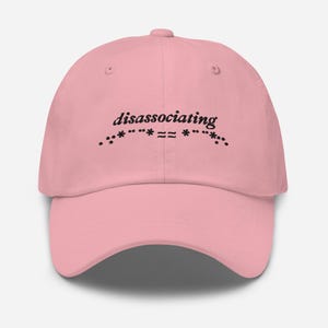 Op de afbeelding: Een roze baseballpet met de tekst "disassociating" geborduurd in het zwart op de voorkant. De tekst wordt omringd door een rij sterretjes.