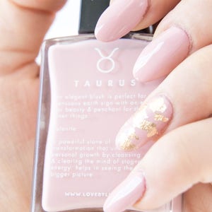 May include: A clear glass bottle of light pink nail polish with a gold Taurus symbol on the label. The bottle is labeled "Taurus" and has text about the zodiac sign. The bottle is held by a hand with pink nails decorated with gold flakes.
