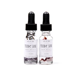 May include: Two glass bottles of Moon Shine gem elixir with black droppers. One bottle contains lavender and vanilla, the other rose and cinnamon. Each bottle has a white label with black text, including "LOVE BY LUNA" and "MOON SHINE".