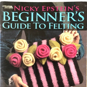 Op de afbeelding: Een boek getiteld "Nicky Epstein's Beginner's Guide to Felting" met een roze en zwart gestreepte gebreide tas met vilten bloemen erop. De tekst op de omslag van het boek luidt "8 Knit Projects for the First-Time Felter! Creative, Boutique-Inspired Designs That Make Knitting Even More Exciting!"