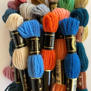 May include: A collection of 16 skeins of DMC embroidery floss in various colors, including teal, orange, blue, green, and beige. The skeins are wrapped in black and gold packaging with the DMC logo and the text "100% Pure Virgin Wool" and "Antimite-Mothproof".
