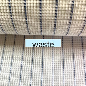 May include: Close-up of a white fabric with thin blue stripes. The fabric is woven in a grid pattern. The word "waste" is printed on a blue label.