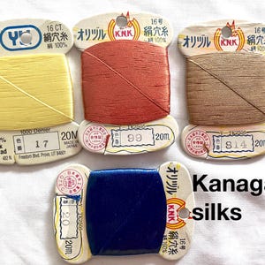 May include: A selection of Kanagawa silk thread spools in yellow, red-orange, brown, and blue. Each spool is labelled with Japanese text and measurements, including 20m of thread. The text "Kanagawa silks" is also visible.