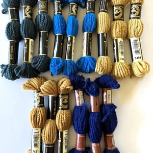 May include: A collection of DMC embroidery floss spools in various colors, including dark green, blue, and gold. The spools are labeled with the DMC numbers 7421, 7494, 7823, and 7317.