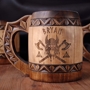 May include: A cylindrical wooden mug featuring a carved geometric border at the top and bottom. The centre is engraved with the name BRYAN above a graphic of a bearded warrior wearing a horned helmet, flanked by two crossed axes and small symbols.