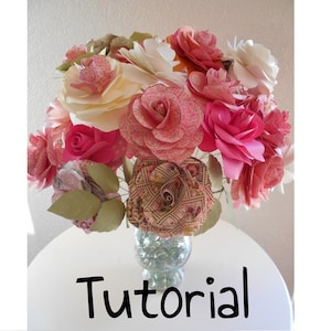 May include: A bouquet of pink and white paper flowers in a clear glass vase with a decorative base. The flowers are arranged in a circular pattern and have a delicate, feminine look. The vase is filled with small, clear glass beads. The word "Tutorial" is written in black letters on a white background below the vase.