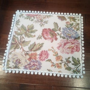 May include: A white square cushion with a floral pattern and a white pom-pom trim.