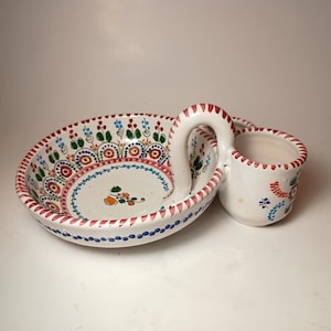 May include: White ceramic serving dish with a handle and a small cup. The dish is decorated with colorful floral and geometric patterns in red, blue, green, and orange. The rim has a red and white striped design.