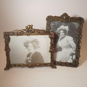 May include: Two ornate, antique-style brass picture frames with intricate floral and leaf relief detailing. The smaller frame is landscape-oriented, while the larger frame is portrait-oriented, both containing black and white vintage portraits.