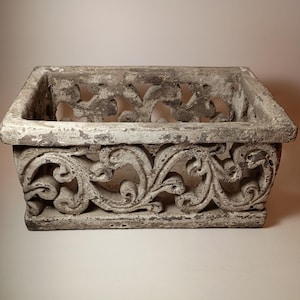 May include: A rectangular, weathered gray planter with ornate, scrolling floral designs. The planter's sides and top edge feature detailed carvings, creating a vintage aesthetic. The distressed finish adds to its antique charm, suitable for garden decor.