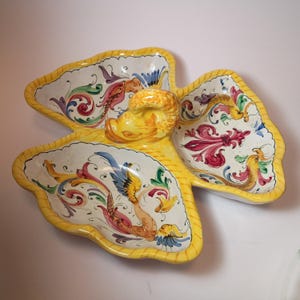 May include: A decorative, hand-painted ceramic serving dish. The dish is shaped like a flower with three petal-shaped compartments and a central, raised element. The dish features a yellow border and is decorated with colorful floral and mythical creature motifs.