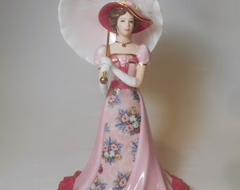 May include: A porcelain figurine of a woman in a pink dress with floral print, holding a white parasol. She is wearing a pink hat with a feather and a pearl necklace.