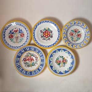 May include: Five hand-painted ceramic plates with floral designs. Each plate features a white center with a different colorful floral arrangement, a blue or yellow border, and a yellow rim. The plates are arranged on a white surface.