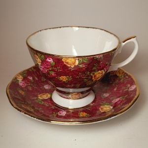 May include: A porcelain teacup and saucer set featuring a deep burgundy background with a repeating pattern of yellow and pink roses. The cup has a white interior, a pedestal base, and a handle, with gold trim along the rims and handle edges.