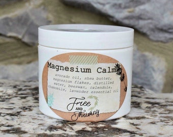 May include: A white jar with a brown label that reads "Magnesium Calm" and lists ingredients including avocado oil, shea butter, magnesium flakes, distilled water, beeswax, calendula, chamomile, and lavender essential oil. The label also includes the text "Free and Thriving".