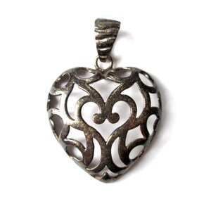 May include: A silver heart-shaped pendant with an intricate, openwork design. The pendant has a delicate, filigree pattern and is perfect for adding a touch of elegance to any outfit.