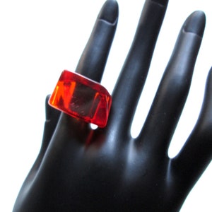 May include: A bright red, rectangular ring made of clear plastic. The ring is worn on a black mannequin hand.