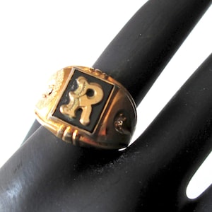 May include: A gold-coloured ring with a black square centre featuring a gold letter "R". The ring has a textured band and decorative details on the sides. The ring is on a black surface.