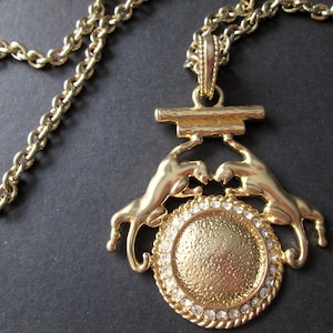 May include: Gold-coloured necklace with a pendant depicting two stylised panther figures holding a textured circular medallion. The medallion is adorned with small, clear crystals. The chain is a classic link style.