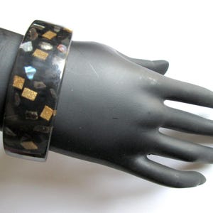 以下が含まれることがあります： 黒い幅広バングルブレスレット。金と虹色の長方形のアクセントが埋め込まれています。白い背景に黒い手で表示されています。このジュエリーは大胆なステートメントピースです。