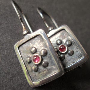 May include: A pair of silver earrings. Each earring has a rectangular shape with a raised border and a central cluster of small, dark grey spheres around a round, red gemstone. They have a hook closure.