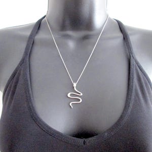 May include: A silver-toned snake pendant suspended from a thin, delicate silver-toned chain. The pendant features a stylised, minimalist serpent silhouette with a smooth, polished finish, hanging centrally against a plain black background.