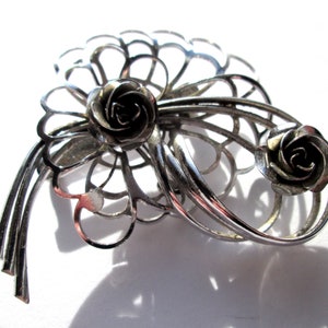 May include: A silver brooch featuring two roses, one larger than the other, set within a delicate, openwork design. The brooch is crafted from silver and has a polished finish.