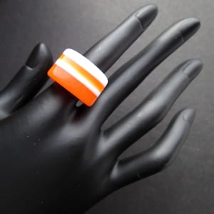 Peut inclure: Bague en verre rayée orange et blanche. La bague est sur une main de mannequin noire.