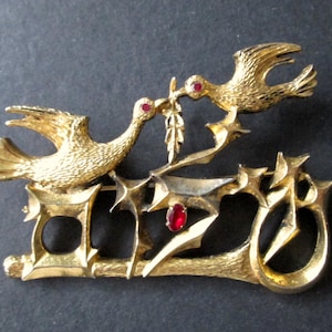 May include: Gold-tone brooch featuring two doves perched on a stylized branch with red rhinestone accents. The doves are facing each other and appear to be sharing a sprig of holly.