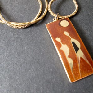 Peut inclure: Un collier à chaîne serpent dorée avec un pendentif rectangulaire en émail orange brûlé brillant. Le pendentif présente un motif stylisé de deux silhouettes abstraites se tenant sous une lune circulaire, réalisé en métal doré poli.
