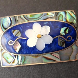 May include: Rectangular silver brooch with an oval blue centre featuring a white flower with a gold centre. The brooch is surrounded by iridescent abalone shell pieces.
