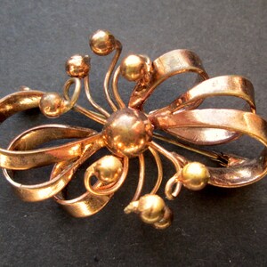 May include: A gold-toned brooch featuring a stylised bow design. The bow is made of multiple loops of metal and has a central round element. The brooch is decorated with small round beads.