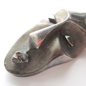 May include: A silver-toned, abstract face sculpture with a textured surface. The sculpture features a prominent nose, eye, and mouth, with a stylized design. The piece has a modern, artistic aesthetic.