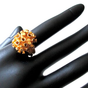 Peut inclure: Bague dorée avec un design texturé et poreux, rappelant une éponge de mer ou du corail. Les détails complexes de la bague sont mis en valeur sur un fond noir. Ce bijou distinctif est une pièce unique, parfaite pour ajouter une touche d'originalité.