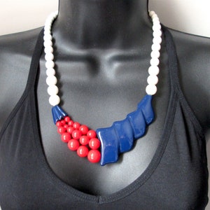 May include: A white, red and blue statement necklace with a large blue plastic piece in the centre. The necklace is made of plastic beads.