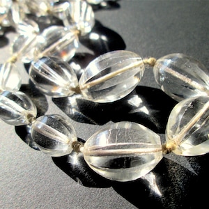 May include: Close-up of a clear glass bead necklace. The oval beads have vertical ridges and a central line. They are strung on a light brown string. The background is a dark grey surface.