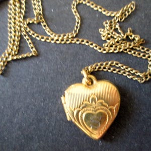 May include: A gold-toned heart-shaped locket pendant suspended from a matching gold-toned link chain. The locket features a textured surface with vertical lines on the upper portion and an engraved decorative pattern surrounding a central heart shape.
