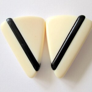 May include: A pair of cream-coloured, triangular earrings with a diagonal black stripe. The earrings have a smooth, glossy finish and a geometric design. The earrings are a vintage style.